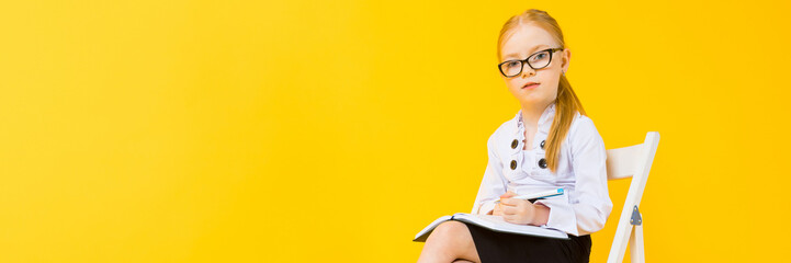 Wall Mural - Girl with red hair on a yellow background. A charming girl in transparent glasses sits on a white chair and makes notes in a notebook.