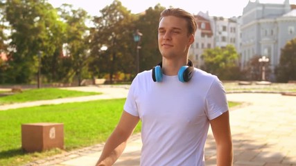 Leinwandbilder - handsome caucasian guy walking in city park. student with headphones wearing white t-shirt strolling slow motion