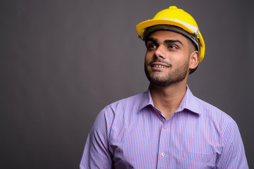 Wall Mural - Young handsome Indian businessman wearing hardhat against gray b