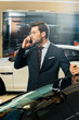 © LIGHTFIELD STUDIOS - smiling young businessman in eyeglasses talking on smartphone near car at dealership salon