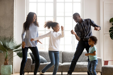 Wall Mural - Happy African American having fun together indoors, funny married couple dancing with adorable little preschooler daughter and cute toddler son at home, listening to music, family weekend with kids