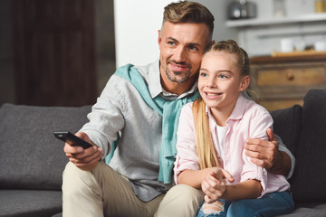 father with remote controller sitting on sofa and hugging daughter