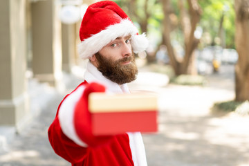 Serious determined guy giving Christmas gifts. Bearded young man wearing Santa Claus costume offering present box at camera. Christmas presents concept