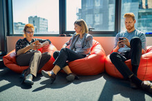 Colleagues With Cell Phones Sitting In Bean Bags In Office Lounge