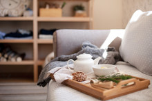 Morning At Home, Tea Time. Wooden Serving Tray With White Teapot, Mug Of Hot Beverage. Cozy Interior In Living Room. Breakfast Is On Sofa With Knitted Sweater In Warm Light. Autumn, Winter Atmosphere.
