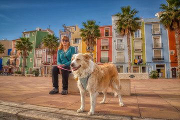 Focus on a blonde border collie mix dog on the boardwalk of Vila Joiosa in Spain with multicolored building and a blonde woman in the background.