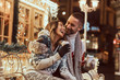 © Fxquadro - A young romantic couple wearing warm clothes outdoor at Christmas time, sitting on a bench and warming with hot coffee in evening street decorated with beautiful lights.