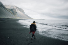 Young Woman In Cool Outdoor Outfir, Red Shirt And Puffy Down Jacket Walk In Empty Cold Grey Beach With Black Volcanic Sand. Concept Explore More And Unconventional Travels In Iceland