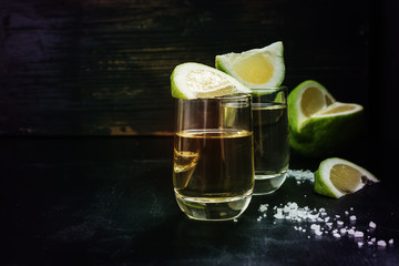 Golden tequila shots with sea salt, juicy lime on vintage black wooden board, toned, selective focus, copy space.