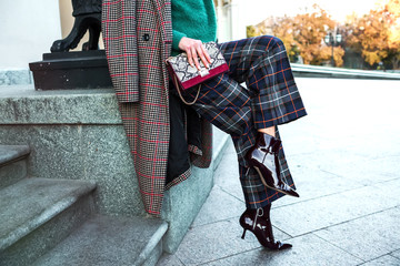 Close up fashion woman sitting in high heel shoes hold black trendy handbag . Stylish outfit checkered pants and coat.Street fashion details of elegant outfit,ankle sock boots, holding leather handbag