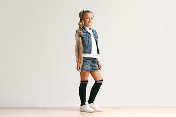 Full length portrait of cute little teen girl in stylish jeans clothes looking at camera and smiling against white studio wall. Kids fashion concept