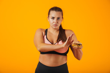 Image of serious chubby woman in tracksuit doing stop gesture while holding sandwich, isolated over yellow background