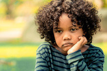 Unhappy Bored Little African American Kid Sitting In The Park. The Boy Showing Negative Emotion. Child Trouble Concept.