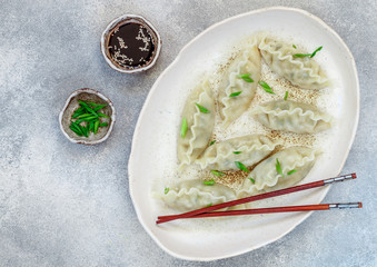 Korean dumplings with meat and vegetables on a white plate with sesame, soy sauce and green onions on a gray concrete surface. Asian cuisine. Selective focus