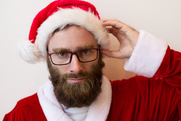 Sticker - Serious bearded man wearing Santa Claus costume and glasses. Handsome young guy looking at camera and touching head. Christmas and business concept. Isolated front view on white background.