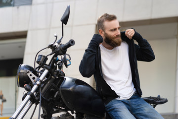 Smiling young biker with motorcycle putting on hoodie. Portrait of Caucasian bearded man sitting on motorbike outdoors. Biker culture concept