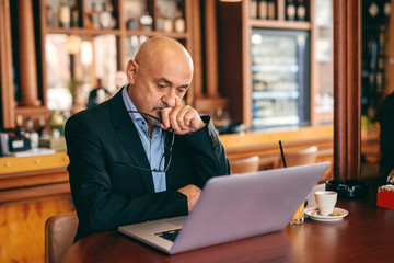 Bearded senior in suit holding hand on his chin while looking at laptop in cafeteria. In hand glasses. On table laptop, coffee and juice.