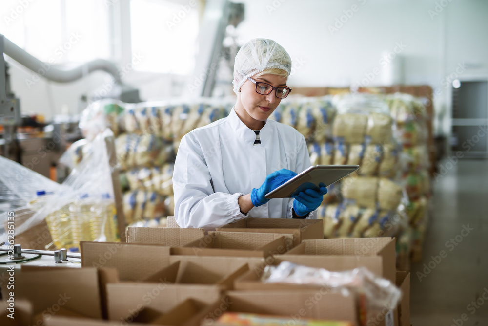 Female worker using tablet for checking boxes while standing in food factory. - obrazy, fototapety, plakaty 