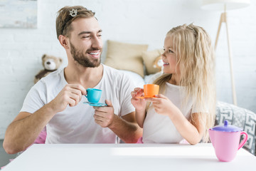 Wall Mural - happy father and daughter smiling each other while playing with toy dishes at home