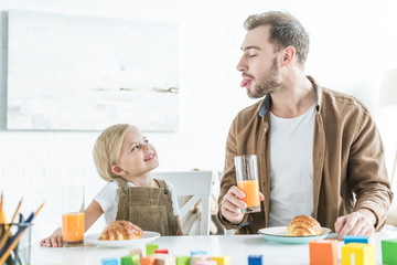 Wall Mural - father showing tongue out to little daughter while having breakfast together