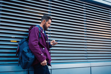 Confident bearded man holding in and mobile phone while standing near street wall in sunny summer evening.