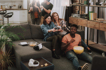 Having fun at home. Joyful girls using joysticks while afro american guy holding popcorn. Lovely couple enjoying beer on background