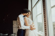 © eduard - Stylish young groom gently hugs his charming brunette bride.Portrait of a young happy couple near the window on black background.
