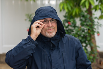 Portrait of senior man with glasses outside in the rain wearing hood and rain coat