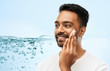© Syda Productions - grooming, skin care and people concept - smiling young indian man applying cream to face over blue background with air bubbles or water splash
