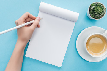 Wall Mural - cropped view of person writing in blank notebook with cup of coffee on blue background