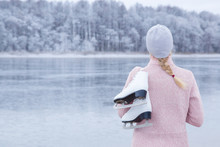 Young Blond Woman In Pink Sweater And Hat Staring At Ice Of Lake And Holding White Skates Over Shoulder In Freezing Winter Day. Back View Of Ice Skater. Outdoor Activities On Weekends In Cold Weather.