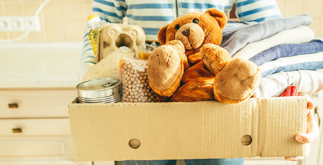 Donating concept - female holding food, clothes, toys in cardboard, kitchen background