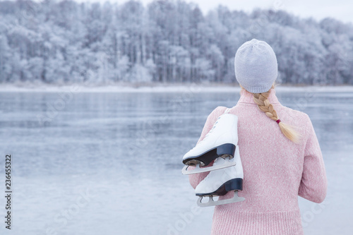 Cuadros en Lienzo Young blond woman in pink sweater and hat staring at ice of lake and holding white skates over shoulder in freezing winter day