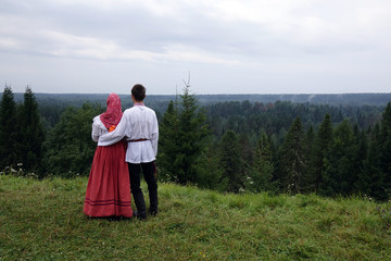 girl and boy in traditional folk costumes in nature, looking into the distance. green pine forest.