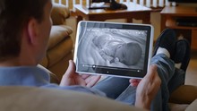 A Father At Home In His Living Room Watches A Baby Monitor On His Tablet PC. Black And White Nightvision Video Of A Sleeping Infant On The Screen.  	