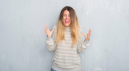 Young adult woman over grunge grey wall wearing winter sweater celebrating mad and crazy for success with arms raised and closed eyes screaming excited. Winner concept