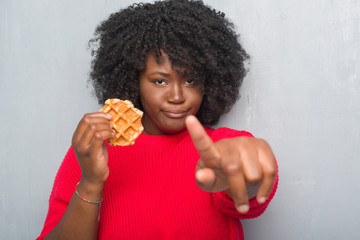 Canvas Print - Young african american woman over grey grunge wall eating belgium waffle pointing with finger to the camera and to you, hand sign, positive and confident gesture from the front