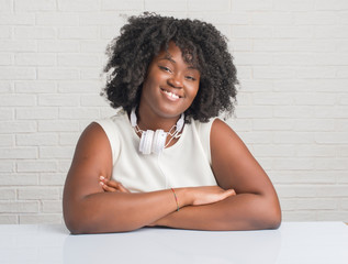 Poster - Young african american woman sitting on the table wearing headphones happy face smiling with crossed arms looking at the camera. Positive person.