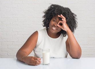 Wall Mural - Young african american woman sitting on the table drinking a glass of milk with happy face smiling doing ok sign with hand on eye looking through fingers