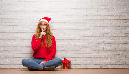 Poster - Young redhead woman sitting over brick wall wearing christmas hat asking to be quiet with finger on lips. Silence and secret concept.