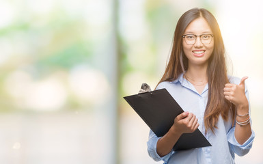 Sticker - Young asian business woman holding clipboard over isolated background happy with big smile doing ok sign, thumb up with fingers, excellent sign