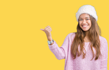 Young beautiful brunette woman wearing sweater and winter hat over isolated background smiling with happy face looking and pointing to the side with thumb up.