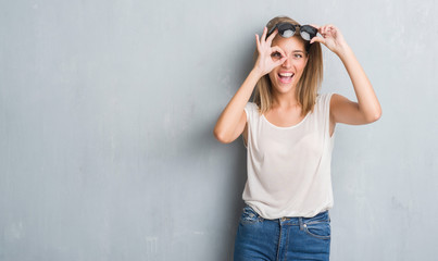 Wall Mural - Beautiful young woman standing over grunge grey wall wearing fashion sunglasses with happy face smiling doing ok sign with hand on eye looking through fingers