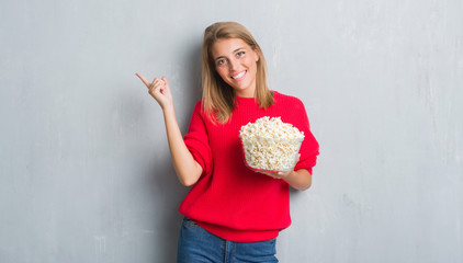 Wall Mural - Beautiful young woman over grunge grey wall eating pop corn very happy pointing with hand and finger to the side