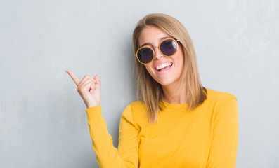 Wall Mural - Beautiful young woman standing over grunge grey wall wearing retro sunglasses very happy pointing with hand and finger to the side