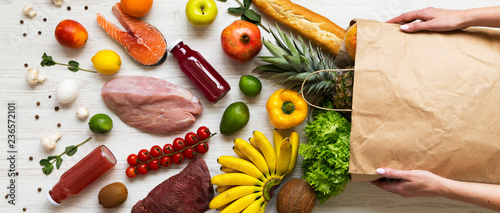 Fotografía Women's hands hold full paper bag of groceries over white wooden background, top view