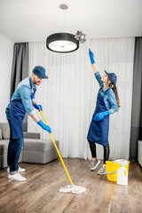 Wall Mural - Professional cleaners in blue uniform washing floor and wiping dust from the lamp in the living room of the apartment. Cleaning service concept