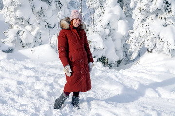 A girl in a pink knitted hat with a pompon and white mittens with a pattern in a warm jacket of copper color with a fur hood stands against the mountain and snow-covered trees