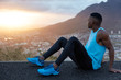 Fitness male sits sideways, has black skin, muscular hands, dressed in sportswear, looks attentively at sunrise, poses over mountains background, takes break after intensive running. Sport, nature