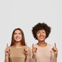 Diverse cheerful women point above, offer to use copy space wisely, smile joyfully, wear casual jumpers, stand against white studio wall, show great place upstairs for shopping and entertainment